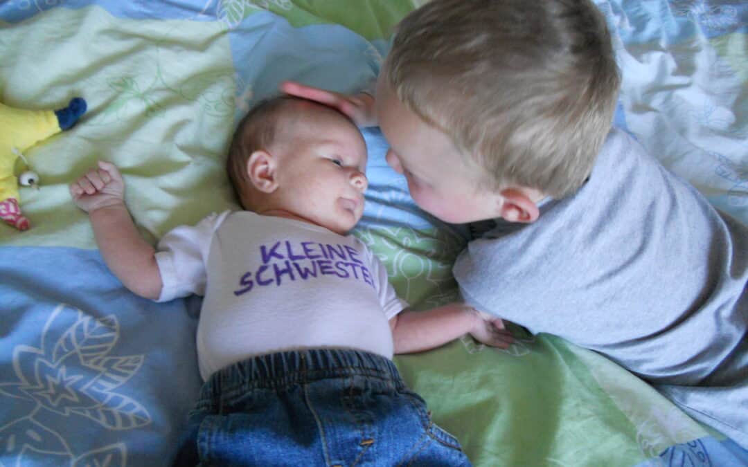 Ein Baby trägt ein Shirt mit der Aufschrift Kleine Schwester. Ein älteres Kind streichelt das Baby am Kopf. Das ist das Bonuskind, der Halbbruder.