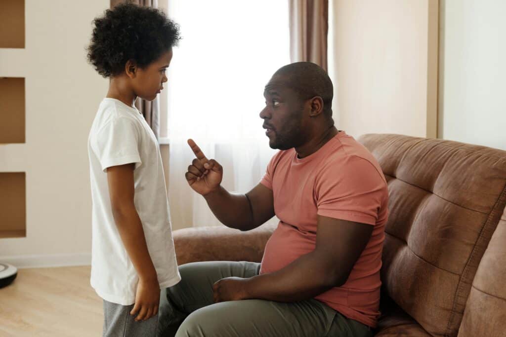Ein Vater sitzt vor seinem stehenden Sohn. Er hat einen erhobenen Zeigefinger und schaut ernst. Ob er mit ihm schimpft?